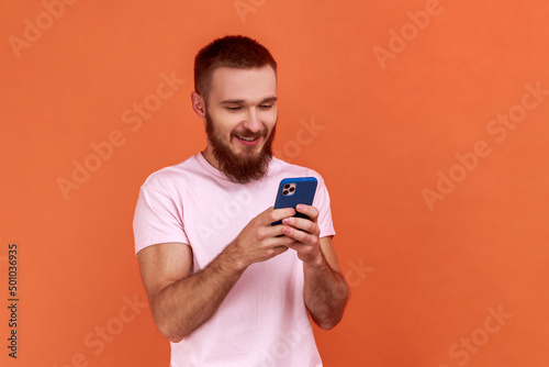 Portrait of bearded man using mobile phone with happy expression, addicted to smartphone, texting in social network, wearing pink T-shirt. Indoor studio shot isolated on orange background. © khosrork