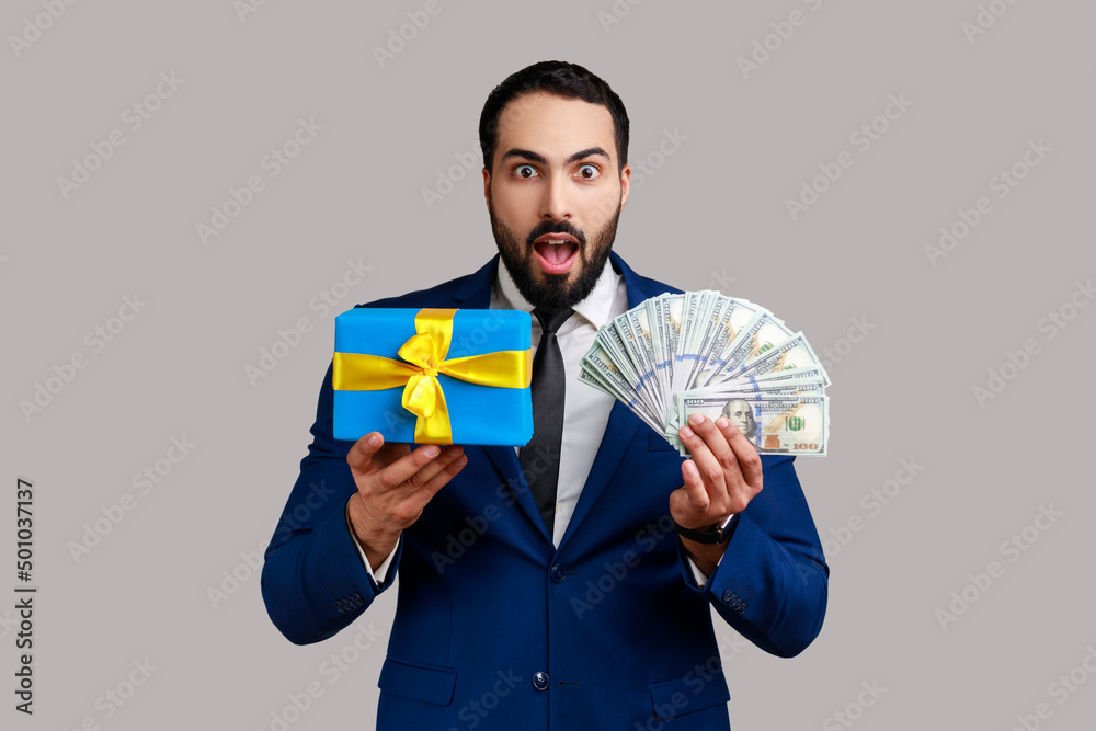 Shocked astonished bearded man holding gift box and dollar money, being surprised excited about cashback, sales, wearing official style suit. Indoor studio shot isolated on gray background. Shocked astonished bearded man holding gift box and dollar money, being surprised excited about cashback, sales, wearing official style suit. Indoor studio shot isolated on gray background.