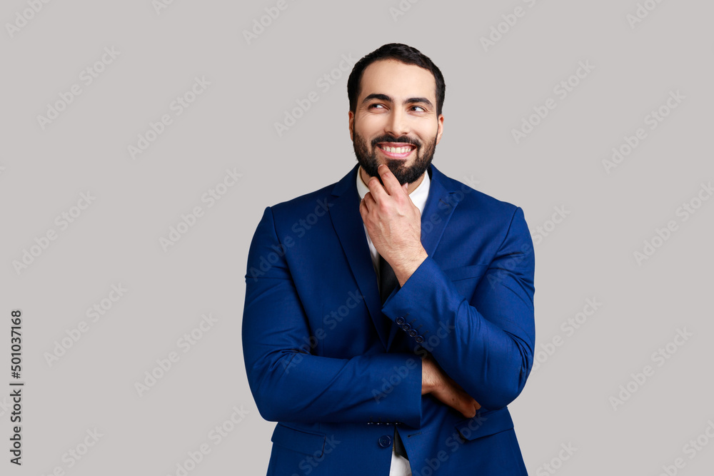 Bearded man with peaceful face thinking over plans for future holding his chin, thoughtful male dreaming about good job, wearing official style suit. Indoor studio shot isolated on gray background. Bearded man with peaceful face thinking over plans for future holding his chin, thoughtful male dreaming about good job, wearing official style suit. Indoor studio shot isolated on gray background.