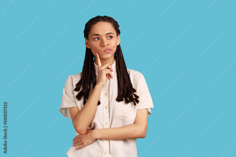 Portrait of thoughtful woman with black dreadlocks thinking about future, holding chin, having serious facial expression, wearing white shirt. Indoor studio shot isolated on blue background. Portrait of thoughtful woman with black dreadlocks thinking about future, holding chin, having serious facial expression, wearing white shirt. Indoor studio shot isolated on blue background.