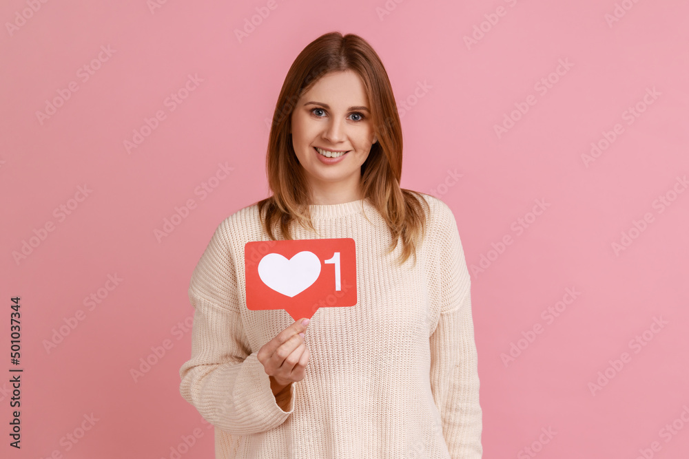 Portrait of blond woman holding social media heart Like icon and smiling to camera, recommending to follow and love content, wearing white sweater. Indoor studio shot isolated on pink background. Portrait of blond woman holding social media heart Like icon and smiling to camera, recommending to follow and love content, wearing white sweater. Indoor studio shot isolated on pink background.