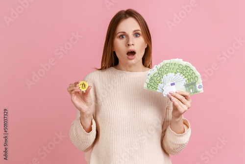 Astonished surprised woman holding gold coin of crypto currency and big fan of euro banknotes, looking at camera with open mouth, wearing white sweater. Indoor studio shot isolated on pink background. photo