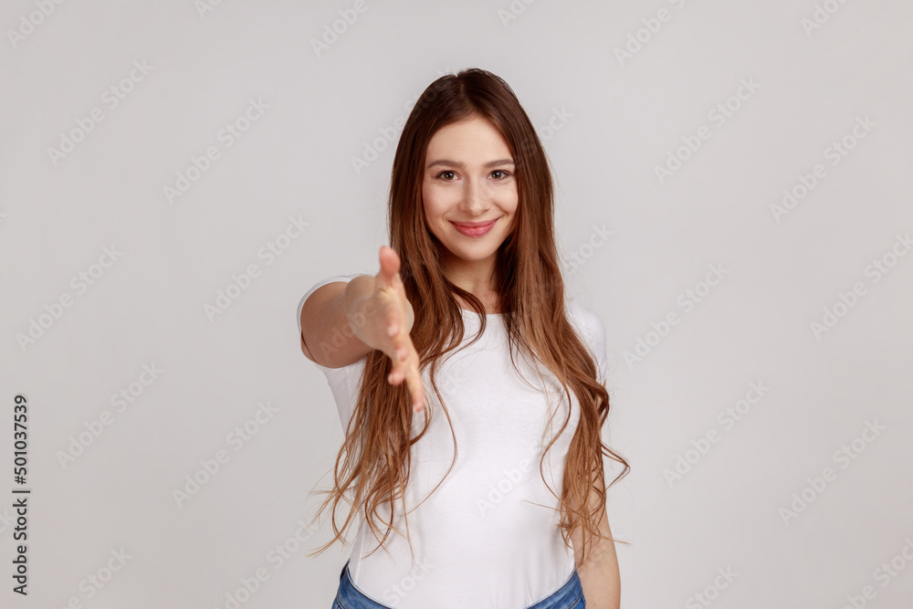 Let me introduce myself. Portrait of friendly woman giving hand to handshake, greeting guests with toothy smile, wearing white T-shirt. Indoor studio shot isolated on gray background. Let me introduce myself. Portrait of friendly woman giving hand to handshake, greeting guests with toothy smile, wearing white T-shirt. Indoor studio shot isolated on gray background.