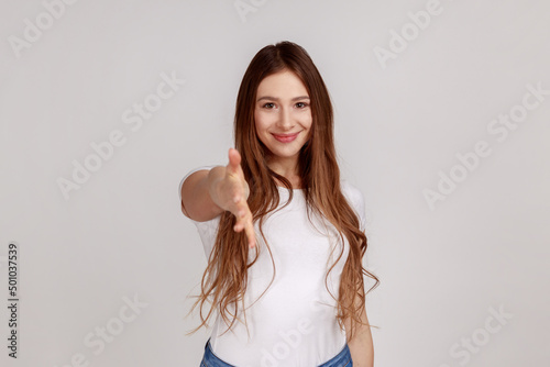 Let me introduce myself. Portrait of friendly woman giving hand to handshake, greeting guests with toothy smile, wearing white T-shirt. Indoor studio shot isolated on gray background.