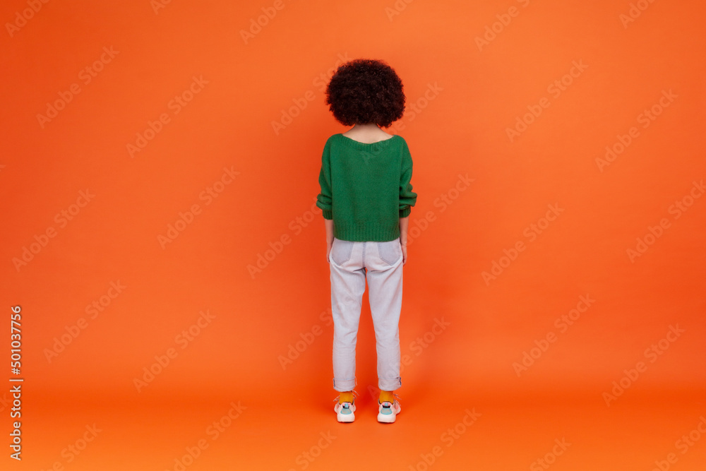 Back view of slim woman with Afro hairstyle wearing green casual style sweater and jeans standing and looking at something, waiting. Indoor studio shot isolated on orange background. Back view of slim woman with Afro hairstyle wearing green casual style sweater and jeans standing and looking at something, waiting. Indoor studio shot isolated on orange background.