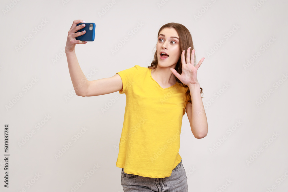 Portrait of happy curly-haired teen girl in yellow T-shirt taking selfie, having conversation on video call and waving hello gesture, vlog broadcast. Indoor studio shot isolated on gray background. Portrait of happy curly-haired teen girl in yellow T-shirt taking selfie, having conversation on video call and waving hello gesture, vlog broadcast. Indoor studio shot isolated on gray background.