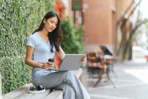 Portrait of beautiful Asian woman sitting outdoors at coffee shop restaurant during summer, using smart wireless technology computer laptop and smartphone, relaxing coffee break at cafe restaurant. © PaeGAG