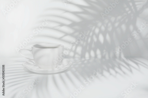 White coffee cup on white table and over white wall with palm leaves shadow background.
