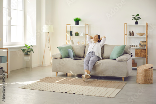 Happy lady leaning back on comfy sofa in cozy bright living room in her own apartment. Relaxed woman enjoying free time on quiet weekend or day off and sitting hands behind head on soft couch at home