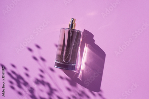 Transparent bottle of perfume on a lilac background. Fragrance presentation with daylight. Trending concept in natural materials with plant shadow. Women's and men's essence.