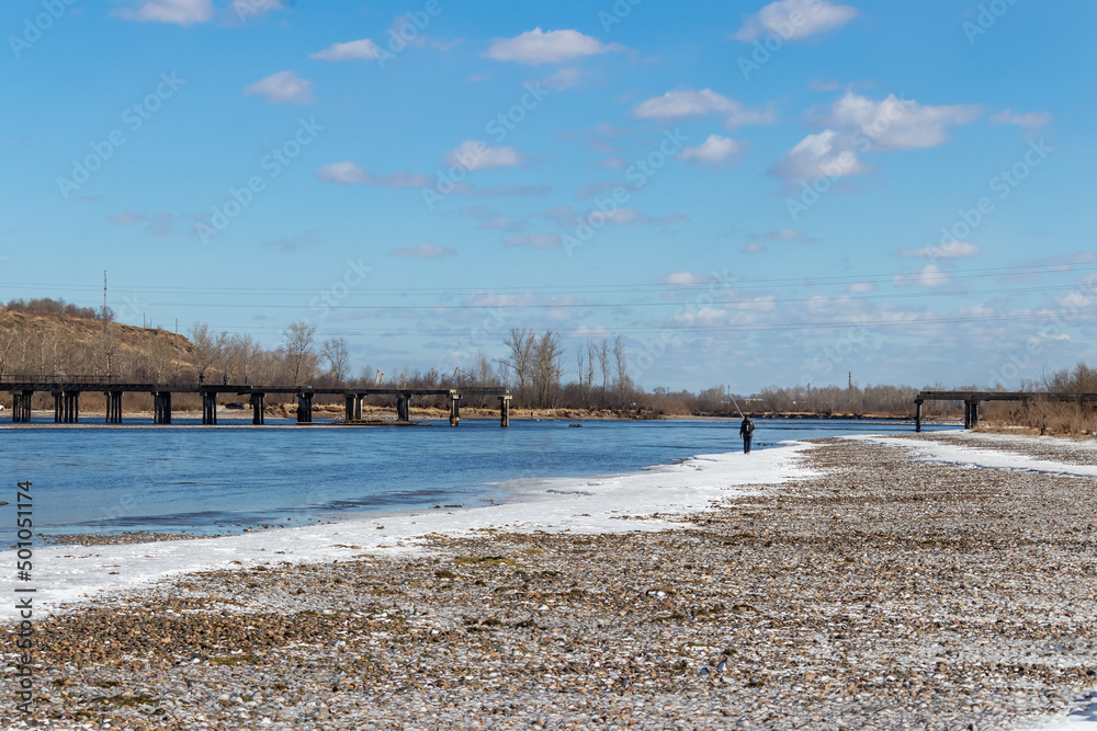 The bank of the Yenisei River against the background of a destroyed bridge, a fisherman walks along the shore on a clear spring day