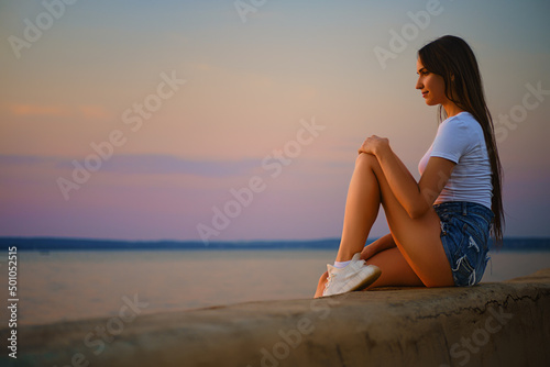 Young pensive woman sitting on a pier and watching the sunset