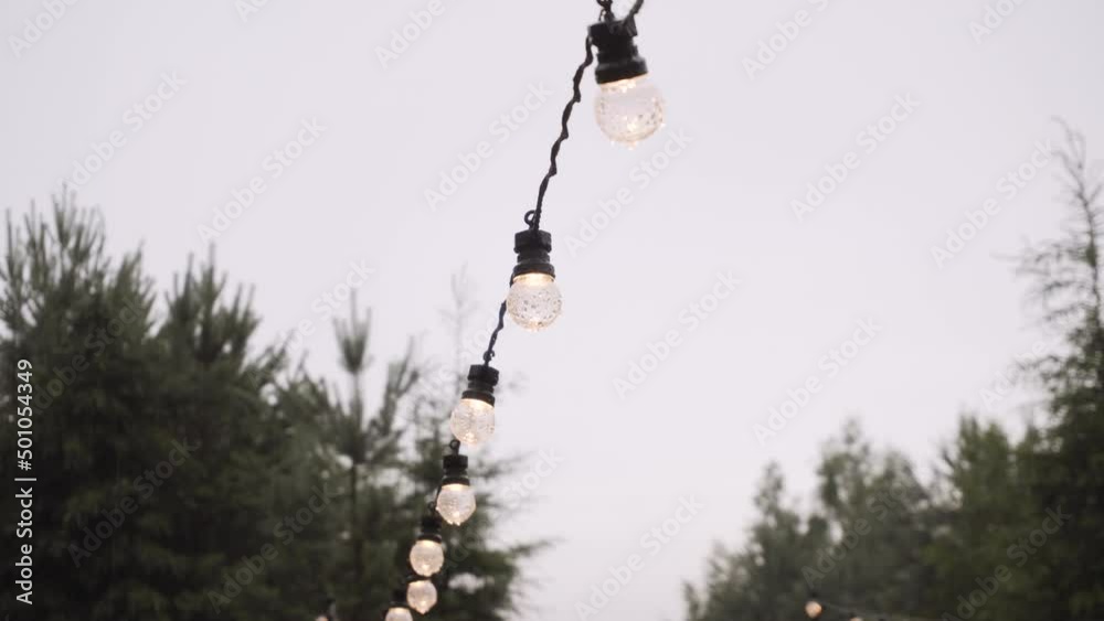 Switched on LED waterproof garland with imitation of glass incandescent bulbs is stretched on site near coniferous trees. Gray sky after rain, bulbs of light bulbs in drops of water. Switched on LED waterproof garland with imitation of glass incandescent bulbs is stretched on site near coniferous trees. Gray sky after rain, bulbs of light bulbs in drops of water.