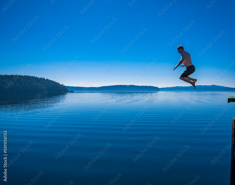 Adventurous athletic man jumping off a pier into a body of calm water.