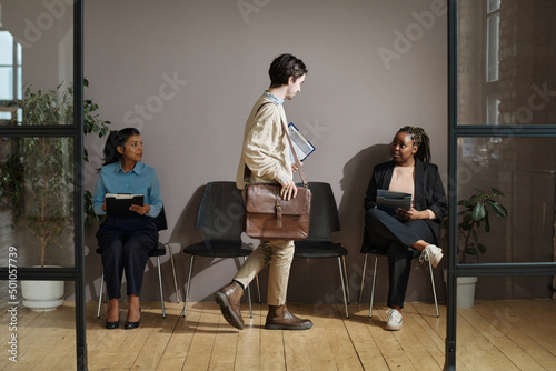 Young man with suitcase sitting on chair between two women in office corrifor