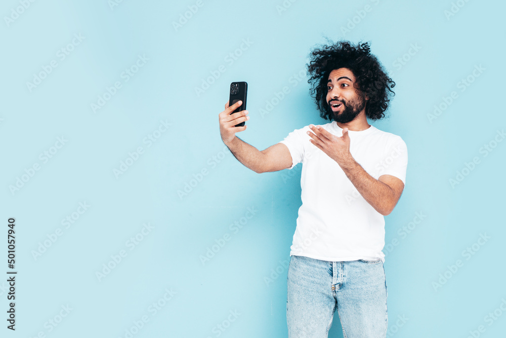 Handsome smiling hipster model.Unshaven Arabian man holding smartphone. Fashion male with long curly hairstyle posing near blue wall in studio. Talking at video call Handsome smiling hipster model.Unshaven Arabian man holding smartphone. Fashion male with long curly hairstyle posing near blue wall in studio. Talking at video call