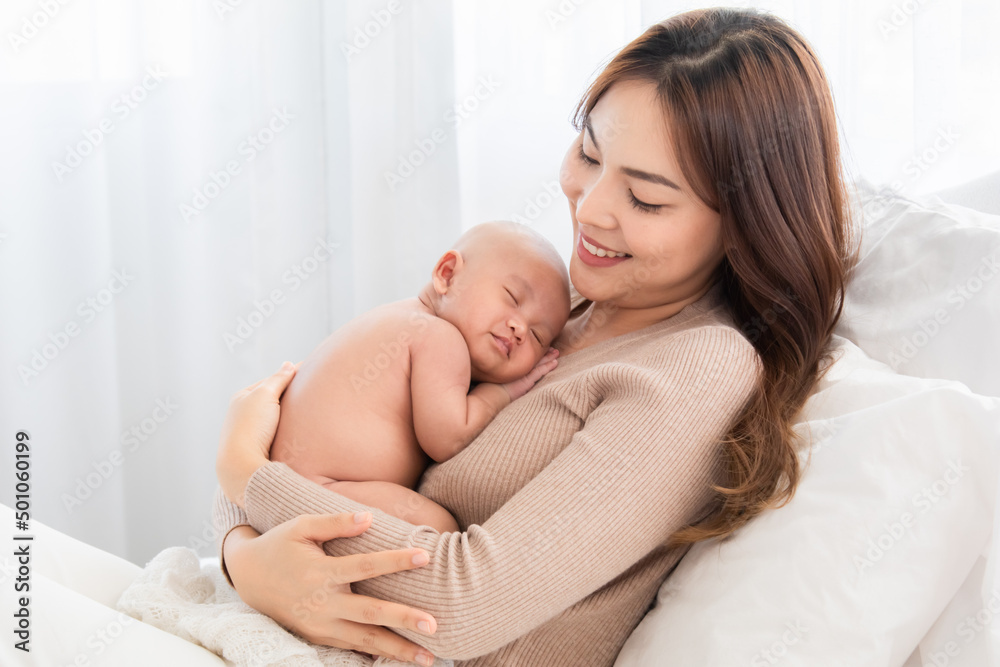 Asian young mother holding tenderly cuddles newborn baby in arms on to chest. Mother looking adorable infant with love and care. Woman and small child lying on bed together at home. Asian young mother holding tenderly cuddles newborn baby in arms on to chest. Mother looking adorable infant with love and care. Woman and small child lying on bed together at home.