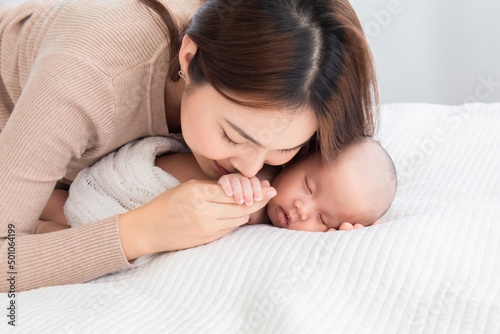 Selective focus beautiful Asian mother kiss hand newborm baby with love and care. Woman hold tiny finger adorable infant while sleeping comfortable on bed at home. Mom and toddler spend time together.