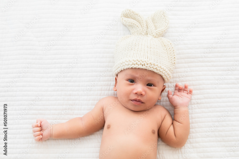 Asian adorable newborn baby wearing pants and rabbit hat beige knitted lying on white bed. little baby 0-1month looking surprised at camera. concept of happy childhood and motherhood. Asian adorable newborn baby wearing pants and rabbit hat beige knitted lying on white bed. little baby 0-1month looking surprised at camera. concept of happy childhood and motherhood.