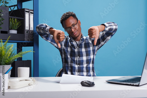 Dissatisfied african american agency manager making disapproval gesture because of unfit marketing strategy of startup project. Finance department displeased team leader giving thumbs down to employee photo