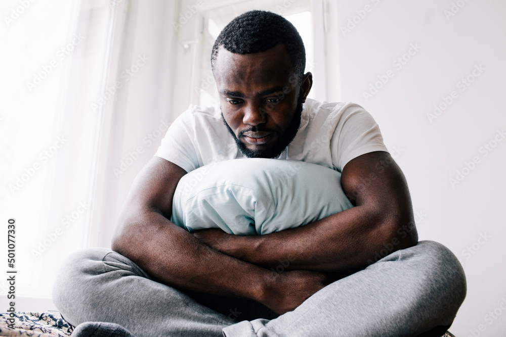 Emotionally stressed man sitting cross-legged with pillow in bedroom at home Emotionally stressed man sitting cross-legged with pillow in bedroom at home