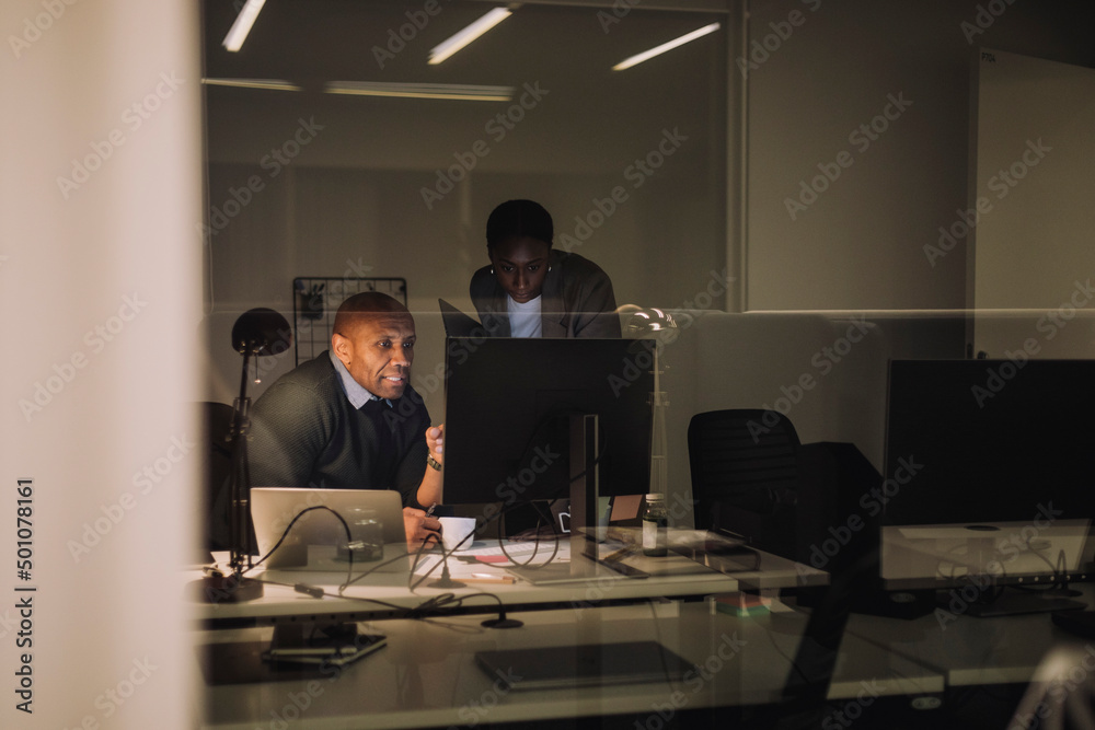 Businessman discussing with female colleague over computer seen through glass at work place Businessman discussing with female colleague over computer seen through glass at work place
