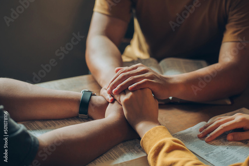Christian family sitting around a wooden table with open bible page and holding hands to bless and pray for each other. comforting and praying together.Christians and Bible study concept.
