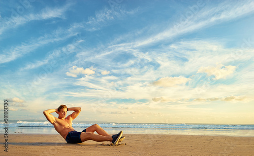Sports and healthy lifestyle. Young man doing crunches on the ocean beach. Enjoying sunset.