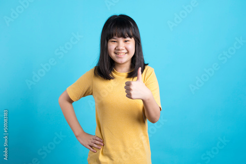 Asian girl gives a thumbs up and smile on blue background.