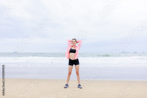 asian woman wearing sportwear and pink jacket exercise and relax on the sea beach at evening in the rain season