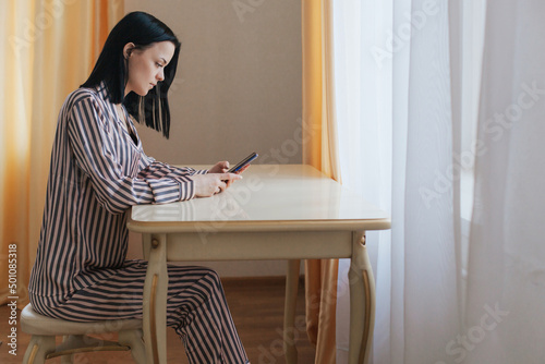 Photo of a young attractive woman in pajamas sitting indoors at a table in a pleasant morning home environment with a mobile phone in her hands.