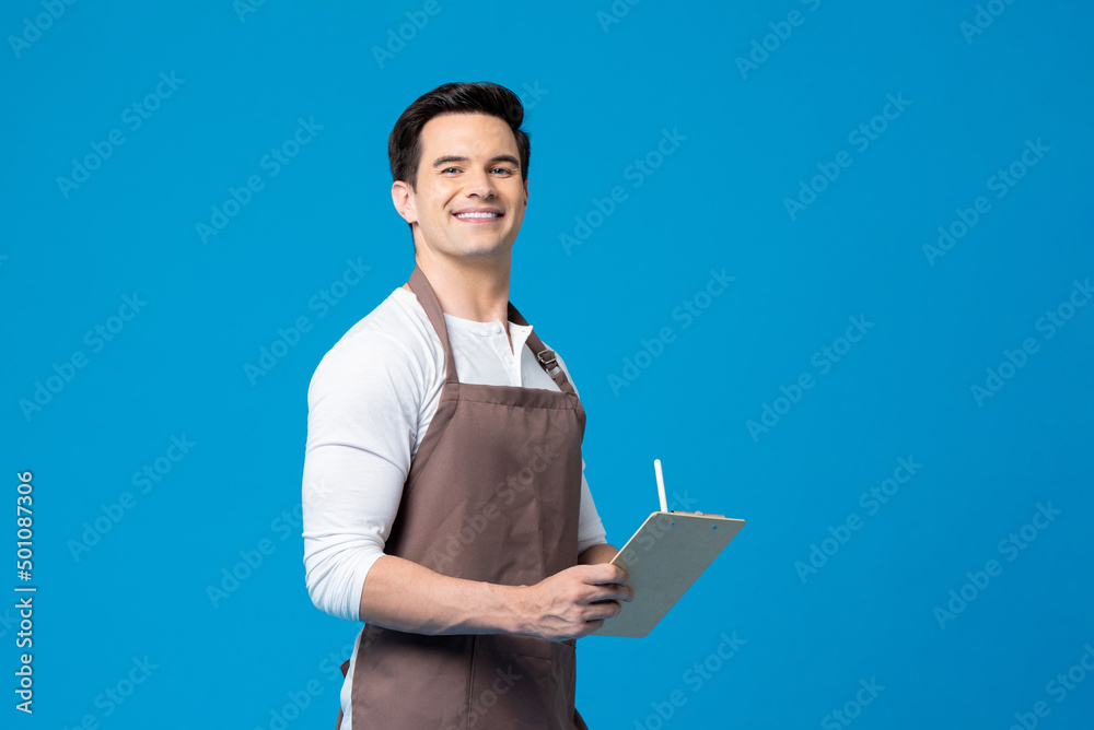 Caucasian man with apron holding writing pad and pen in his hands while standing on blue background in light studio Caucasian man with apron holding writing pad and pen in his hands while standing on blue background in light studio
