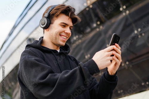 Young Caucasian Man In Headphones Using Smartphone Texting And Listening To Music.