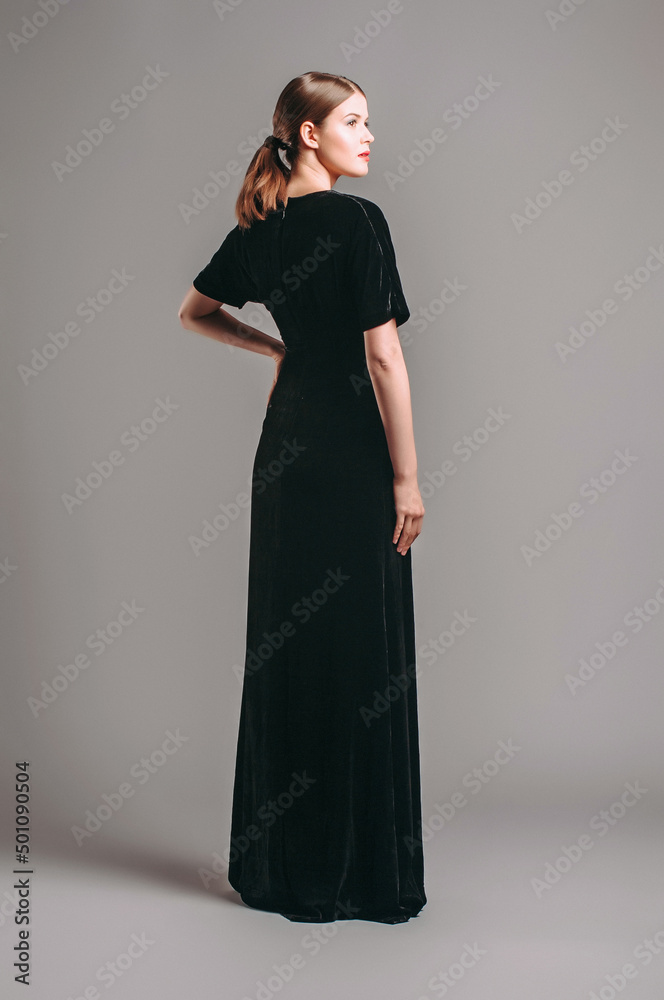 Black velvet trumpet dress. Evening floor length gown with deep v neck line and short sleeves. Beautiful young brunette lady with red lips and ponytail hairstyle posing in studio. Total black look.