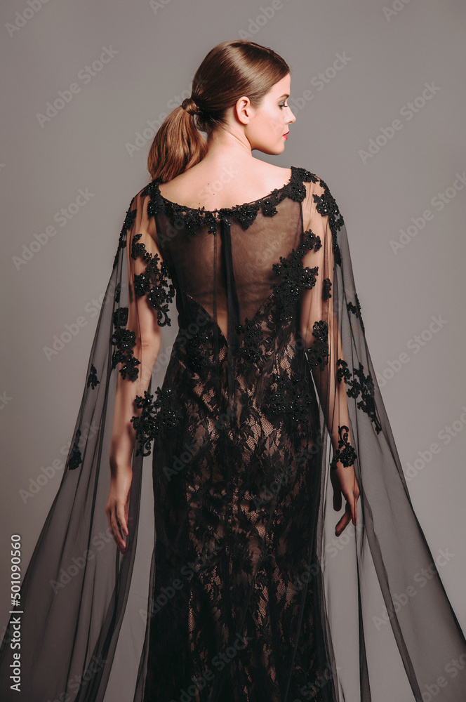 Luxury lace evening gown in black with transparent flutter cape.  Embroidered maxi dress with a train, modern fashion. Fishtail fitted dress.  Brunette model posing in studio. Total black female look. Stock Photo