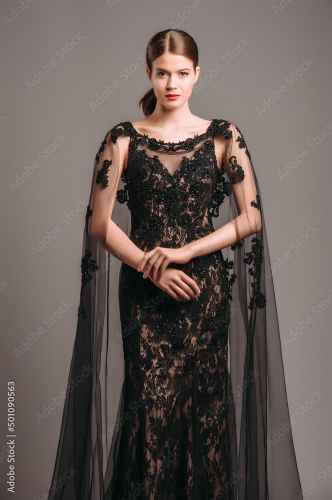 Luxury lace evening gown in black with transparent flutter cape. Embroidered maxi dress with a train, modern fashion. Fishtail fitted dress. Brunette model posing in studio. Total black female look. Luxury lace evening gown in black with transparent flutter cape. Embroidered maxi dress with a train, modern fashion. Fishtail fitted dress. Brunette model posing in studio. Total black female look.