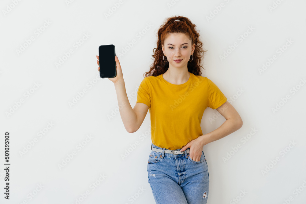 Young happy woman pointing finger at mobile phone screen and smiling pleased, recommending application, useful app on white background Young happy woman pointing finger at mobile phone screen and smiling pleased, recommending application, useful app on white background