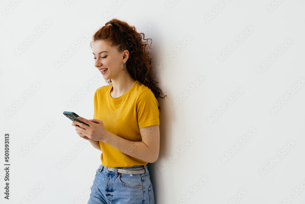 Smiling female student with ginger hair using mobile phone, looking happy at camera, standing over white background. Smiling female student with ginger hair using mobile phone, looking happy at camera, standing over white background.
