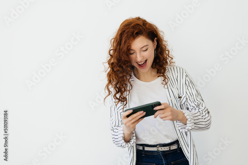 Young redhead woman playing video games on her phone, watching video live stream on cellphone, laughing and smiling, looking at mobile screen, white background
