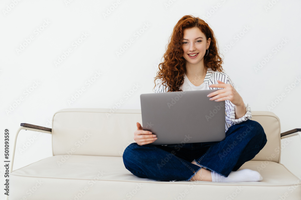 Beautiful red-haired woman working online, using laptop, sitting on comfortable grey couch Beautiful red-haired woman working online, using laptop, sitting on comfortable grey couch