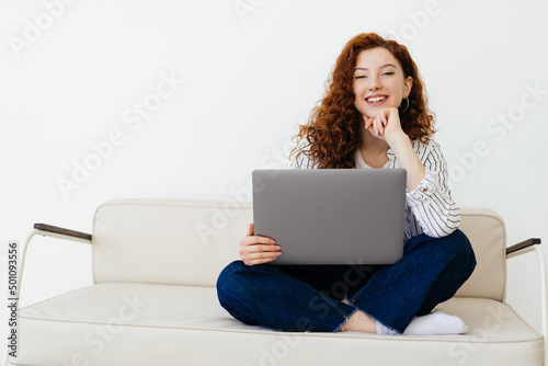 Beautiful young red-haired woman sitting on sofa at home, typing on laptop