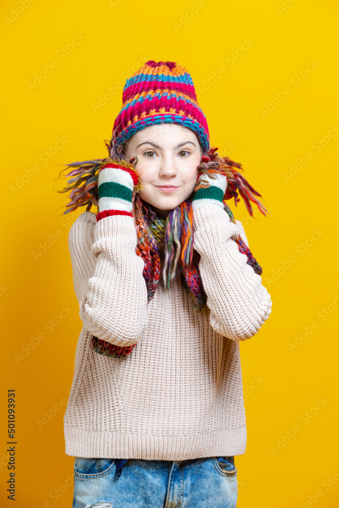 Winsome Caucasian Blond Girl In Warm Knitted Hat and Scarf Posing with Lifted Hands While Touching Cheeks With Knitted Color Scarf Against Yellow. Winsome Caucasian Blond Girl In Warm Knitted Hat and Scarf Posing with Lifted Hands While Touching Cheeks With Knitted Color Scarf Against Yellow.
