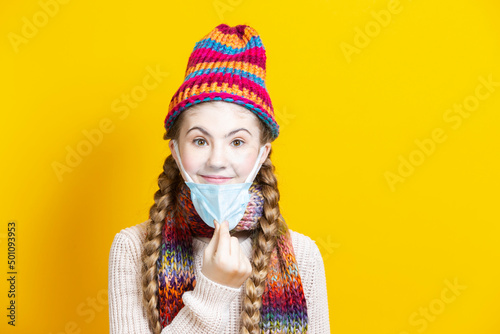 Teenage Girl in Warm Knitted Hat and Scarf Using Facial Mask Against Viruses While Posing Over Trendy Yellow © danmorgan12