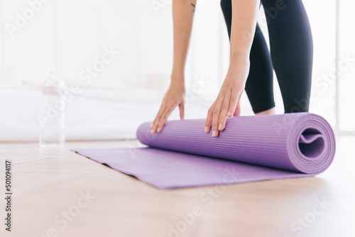Crop image of young woman wearing sportswear rolling yoga mat with water bottle, preparing for doing yoga. Working out at home or in yoga studio. Healthy habits, keep fit, weight loss concepts.