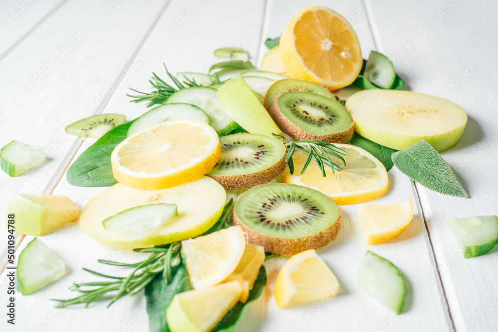 Partially blurred green and yellow fruits and herbs cut into pieces lie on white table. Ingredients for smoothie, fruit salad or dessert. Slices of lemon, apple, kiwi, cucumber, spinach and tarragon