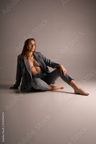 An incredibly beautiful model girl with long blonde hair in an unbuttoned gray jacket, beige bra and gray short trousers is sitting on a floor barefoot. Studio boudoir shooting on a white background