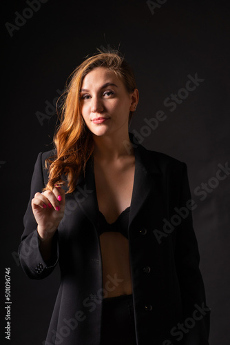 Studio portrait of a gorgeous half-naked smiling sexy blonde in sexy underwear and unbuttoned black jacket on a black background in the semi-darkness playing with her hair (Rembrandt light)