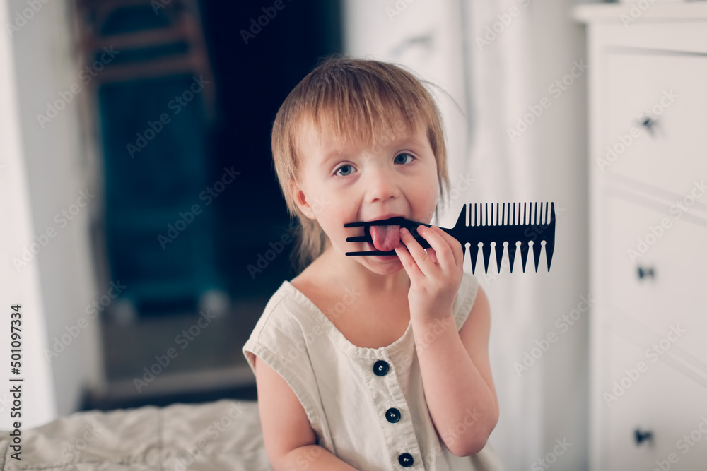 Funny baby toddler in baby overalls nibbles on a comb in bedroom, child safety and teething, dirt and germs Funny baby toddler in baby overalls nibbles on a comb in bedroom, child safety and teething, dirt and germs