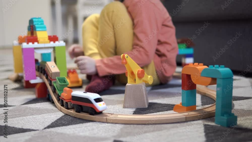 A little boy is playing with a children's railway. The child plays with toys on the floor. Railway bridge from a children's designer. A toy train rides on a wooden railroad.
