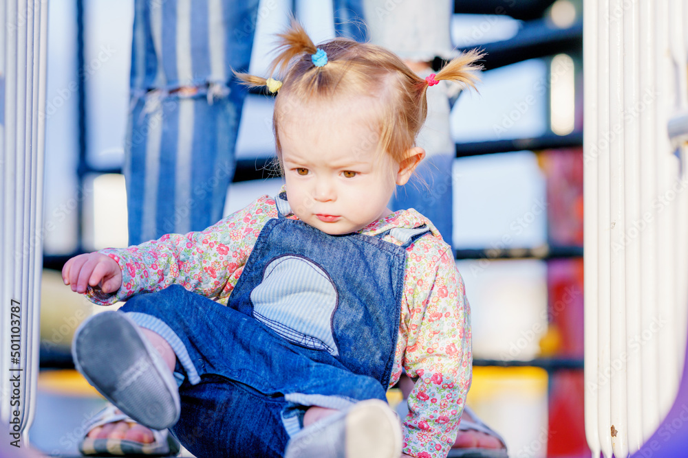 Fototapeta premium Happy child is playing on the playground. Funny girl with pigtails on a children's slide. Baby has multi-colored short pigtails. The kid is learning to climb stairs. Active games for children